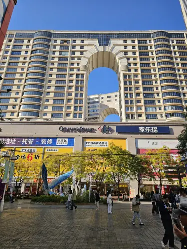 Large residential and commercial building in Kunming with a prominent central arch above a shopping plaza, formerly anchored by a Carrefour hypermarket, photographed on a sunny day with people in the square.