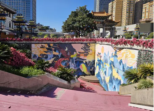 Former entrance to a sunken plaza at Jinma Biji Square in Kunming, now closed and decorated with colorful murals, flowers, and landscaped steps under bright sunlight.