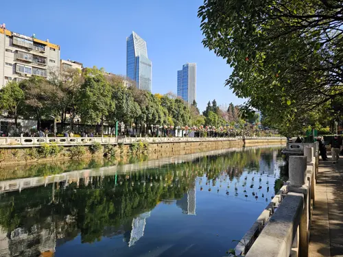 Panlong River in Kunming with clear reflections of a tree-lined walkway and surrounding buildings mirrored in the calm water on a sunny day.