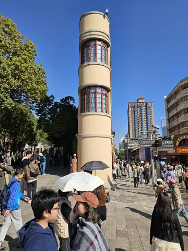 Tall modern building in Kunming seen from its narrow front angle, appearing thin like a vertical slice, photographed on a bright sunny day with people in the surrounding plaza.
