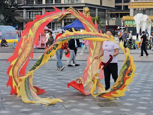 Woman performing with colorful ribbon props in a public square in Jinghong with people around her