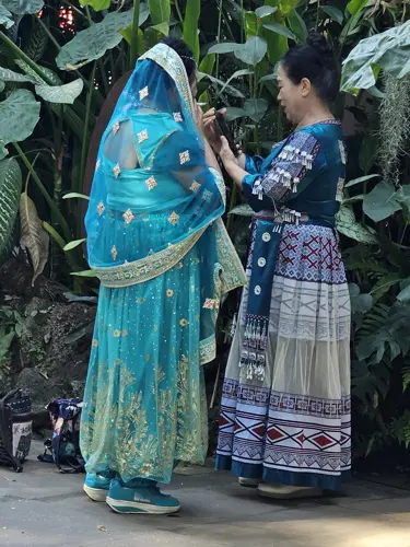 Woman helping another person adjust a traditional-style costume in a leafy outdoor setting in Jinghong