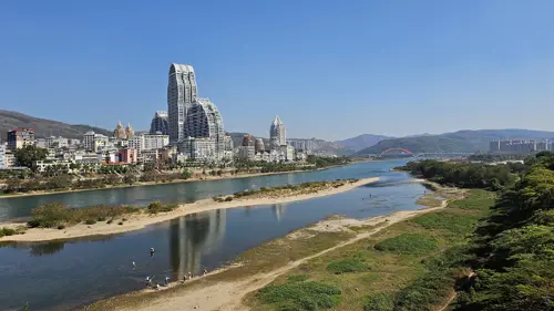 View along the Lancang River in Jinghong with modern buildings and a second bridge visible in the distance