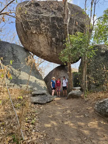Three visitors standing beneath a huge balanced boulder at Wat Khao Hin Thoen